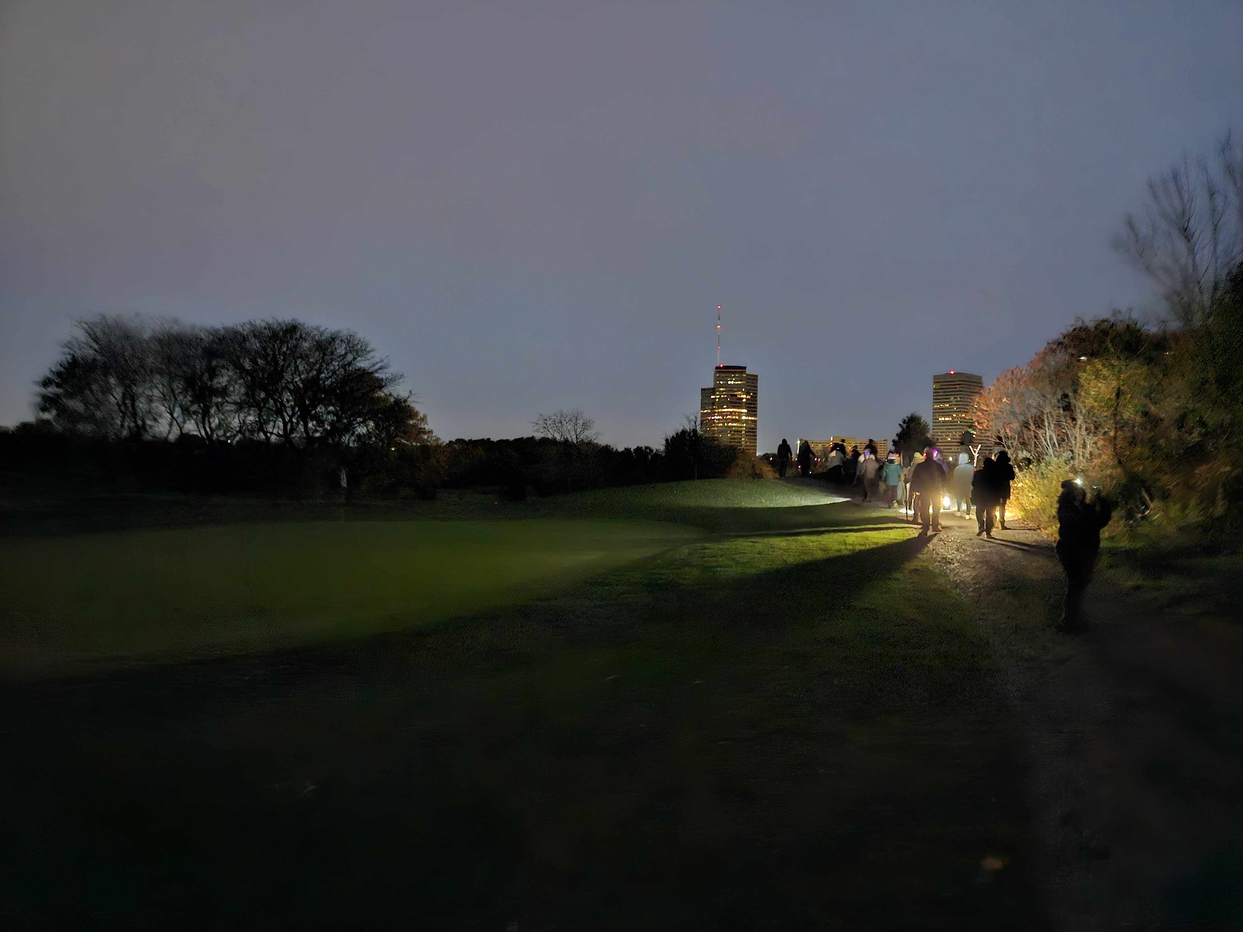 a group of people walking down a path at night illuminated by flashlights, with the Southfield skyscrapers in the background